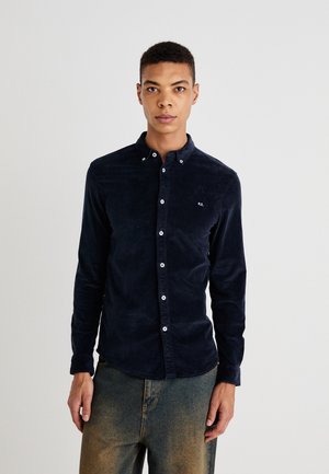 Camisa de pana azul marino con cuello puntiagudo, botones blancos y un sutil detalle de logo en el pecho. Mangas largas con puños de botón.