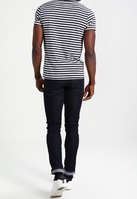 Camisa de manga corta a rayas en azul marino y blanco, combinada con jeans de mezclilla oscuros. Los jeans tienen los puños doblados y zapatillas blancas con acentos en negro.