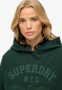 Sudadera verde con el texto "SUPERDAY N.Y.C" en un tono ligeramente más claro, llevada sobre una camisa de cuadros en tonos verdes y marrones.