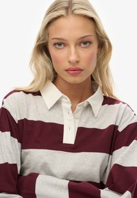 Blonde Frau mit blauen Augen, die ein gestreiftes Rugbyshirt in Weiß und Maroon trägt, schaut direkt in die Kamera mit einem neutralen Ausdruck.