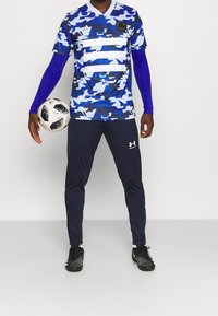 Sininen ja valkoinen kamouflagekuvioinen lyhythihainen paita, yhdistettynä tummansinisiin pitkälahkeisiin housuihin ja mustiin jalkapallokengiin, pitelee jalkapalloa.