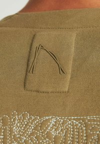Gros plan sur un tissu marron avec un patch rectangulaire cousu et un dessin linéaire simple au-dessus d'un texte blanc brodé sur le textile.