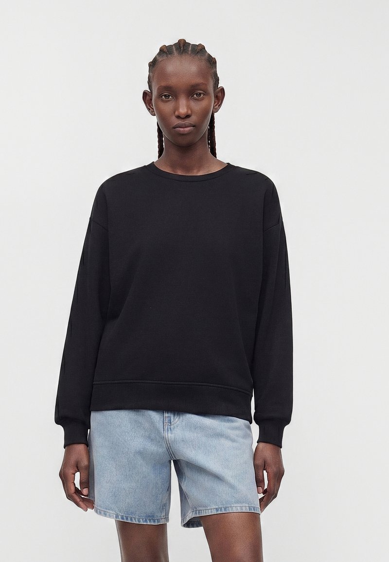 Mujer con el pelo trenzado que lleva una camiseta negra lisa de manga larga y pantalones cortos vaqueros azul claro, de pie frente a un fondo blanco.
