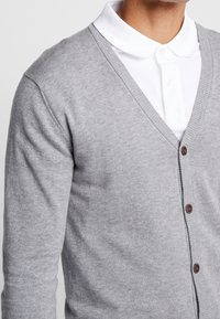Cardigan grigio a maglia con scollo a V, caratterizzato da bottoni marroni e polsini a costine, indossato sopra una polo bianca. Texture morbida, stile aderente.