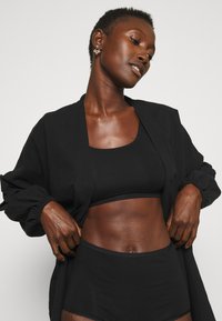 Svart, kort topp og matchende høyt liv shorts, båret under en løs svart cardigan, som fremhever det minimalistiske designet og passformen.
