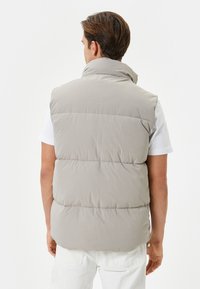 Chaleco acolchado beige claro con cuello alto, textura acolchada y detalle de botón, llevado sobre una camisa blanca, visto desde la parte posterior.