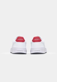 Sneakers bianchi in pelle con accenti rossi sul tallone, dotati di un logo sul tallone e una suola in gomma testurizzata. Design pulito e minimalista.