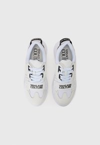 Ténis brancos desportivos com detalhes a preto, com corpo em malha, atacadores, e marca "VERSACE JEANS COUTURE" na lateral e no calcanhar.