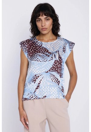 Kobieta o ciemnych falowanych włosach, ubrana w bezrękawnik z abstrakcyjnymi, niebiesko-brązowymi wzorami liści i kropek, w połączeniu z beżowymi spodniami.