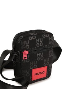 Borsa a tracolla nera in tessuto con un motivo ripetuto "HUGO", cerniera rossa e patch con logo. Forma compatta con tracolla regolabile.