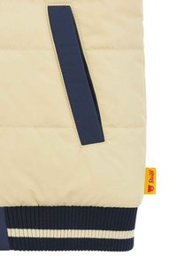 Giacca beige imbottita con accenti blu navy, dotata di una tasca con zip, polsini a coste blu navy e crema, e un'etichetta gialla con logo rosso.