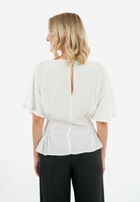 Blusa bianca con maniche corte e fluide e schiena a chiave. Presenta dettagli arricciati in vita, creando un effetto peplum.