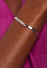 Zilveren armband met een gestructureerd, flexibel ontwerp, gedragen op een donkere arm, tegen een roze textiel achtergrond.