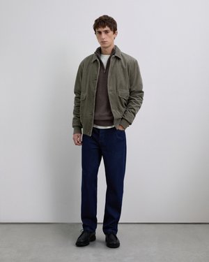 Jonge man leunt tegen een effen muur en draagt een olijfgroene corduroy jas, een bruine pullover, donkerblauwe jeans en zwarte schoenen.