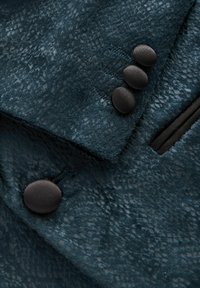 Texturerad, djup teal-färgad tyg med ett subtilt mönster, som har tre svarta knappar och en svart dragkedjeficka på högra sidan.