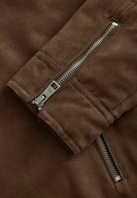 Manica di una giacca in suede marrone con zip metallica argentata parzialmente aperta, che mostra la dettagliata cucitura e la texture del tessuto.