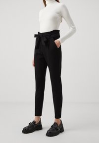 Pantalons noirs ajustés avec un nœud à la taille, dotés de poches latérales et d'une coupe slim, associés à des mocassins noirs à clous massifs et à un col montant en maille blanche.