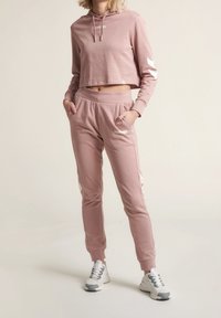 Felpa cropped rosa chiaro con logo bianco, abbinata a jogger coordinati con striscia laterale bianca e vita elastica. Sneakers bianche.