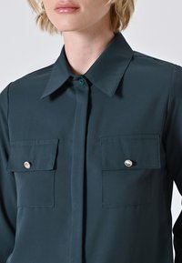 Dunkelblaues T-Shirt mit spitzem Kragen, zwei Brusttaschen mit runden Knöpfen, glatter Textur und Knopfleiste.