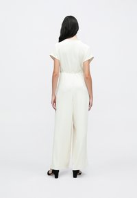 Salopetă albă cu mâneci scurte, talie strânsă și design cu picioare largi. Textură netedă și detaliu cu fermoar în spate, asortată cu pantofi cu toc negri.