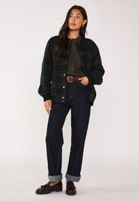 Giacca oversize a scacchi verdi con bottoni a scatto, camicia oliva sotto, jeans scuri arrotolati alle caviglie e mocassini marroni con nappine.