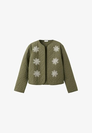 Smilende kvinde iført en olivengrøn quiltet jakke med blomstrede mærker, hvid crop top, jeans med vide ben og sorte sko, stående indendørs.