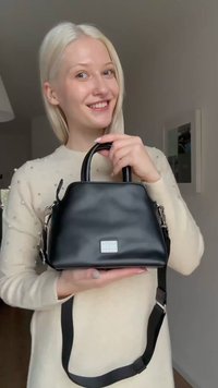 Mlada ženska z dolgimi blond lasmi se smehlja, medtem ko drži črno usnjeno ročno torbico z zgornjimi ročaji in nastavljivim naramnim trakom.