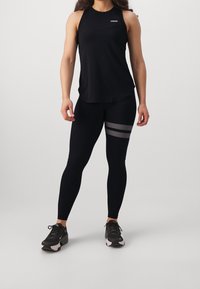 Débardeur de sport noir avec le logo "STRONGER", associé à un legging noir orné d'accents rayés argentés. Le mannequin porte des baskets noires.