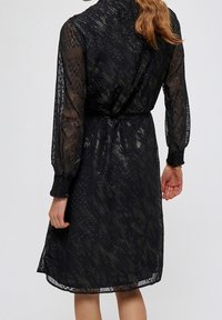 Černé šaty s průhlednými dlouhými rukávy, texturovaným vzorem a staženým pasem. Mají volný střih a délku po kolena.