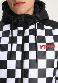 Giacca a scacchi bianca e nera con cappuccio, dotata di logo rosso "YTRN", chiusura con zip e coulisse, realizzata in un materiale liscio e lucido.