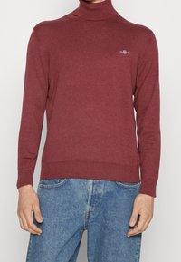 Bordeauxfarbener Rollkragenpullover aus weichem Strickmaterial. Verfügt über lange Ärmel, gerippte Bündchen und Saum sowie ein dezentes Logo auf der Brust.