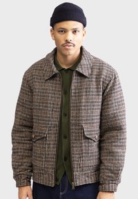 Veste en pied-de-poule marron et crème avec un col chemise, deux poches avant et un ourlet côtelé. Portée par-dessus une chemise à boutons vert foncé.