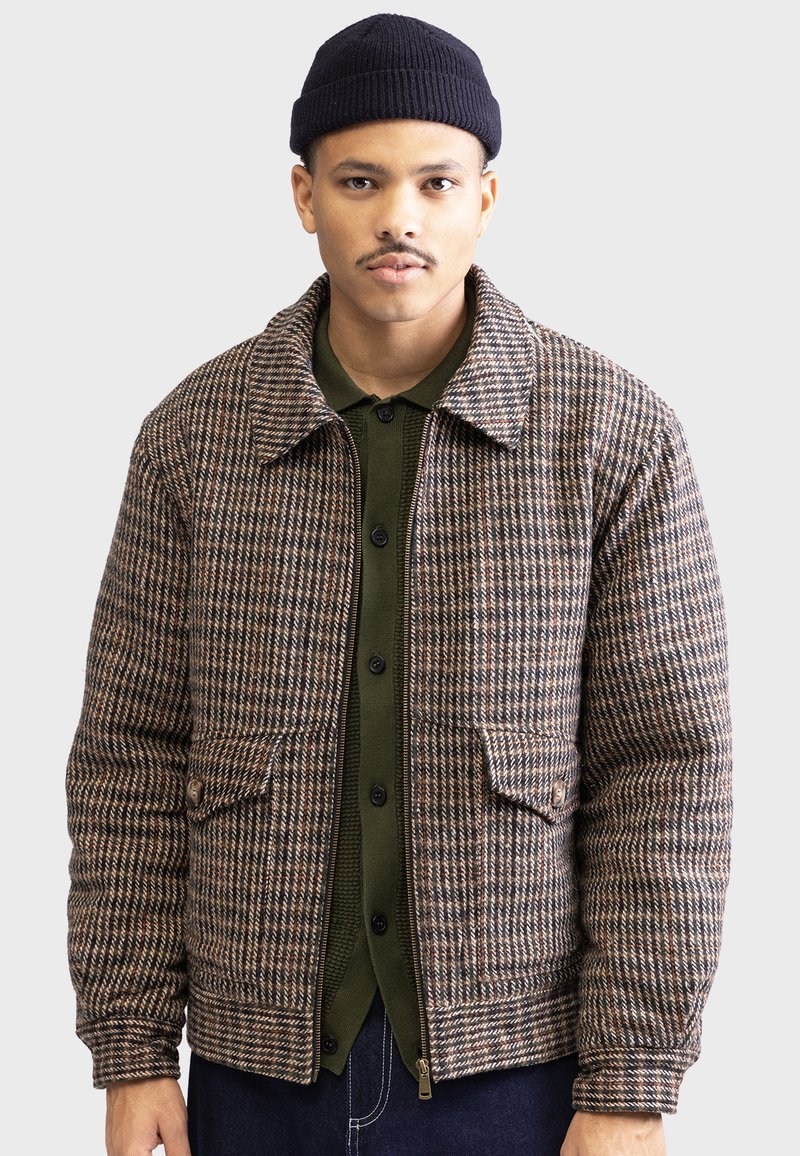 Veste en pied-de-poule marron et crème avec un col chemise, deux poches avant et un ourlet côtelé. Portée par-dessus une chemise à boutons vert foncé.