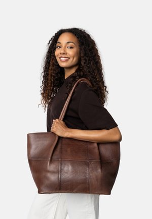 Woman smiling, holding large brown leather tote bag over shoulder, wearing black top and white pants, against plain light background.