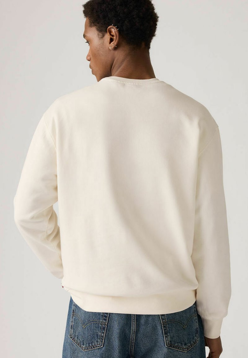 Sweat-shirt à col rond de couleur blanc cassé, avec poignets et ourlet côtelés, texture lisse. Porté avec un jean bleu, présentant des détails de coutures.