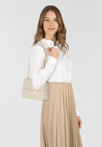 Borsa a tracolla in pelle beige con una cinghia a catena dorata, forma rettangolare, pattina anteriore con chiusura argento e design strutturato. Indossata con una camicia bianca e una gonna beige.