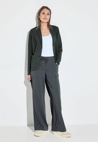 Blazer verde scuro sopra una canotta bianca, abbinato a pantaloni larghi grigi con due tasche frontali e cordoncino, completato da sneakers chiare.