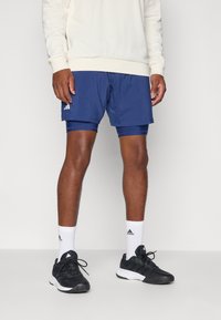 Pantalones cortos deportivos de Adidas en color navy con capa de compresión integrada, combinados con calcetines atléticos blancos y zapatillas negras. Tela suave, ajuste ergonómico.