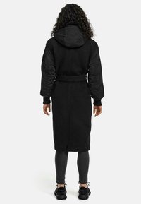 Long black coat with a hood, textured fabric, quilted sleeves, and a waist tie. Worn over dark leggings and casual sneakers.