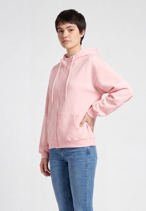 Persoon met kort donker haar, die een lichtroze hoodie met rits en blauwe spijkerbroek draagt, staand met één hand op de heup.