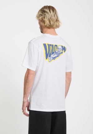Homme blond portant un t-shirt blanc avec un graphisme de squelette en skateboard bleu et jaune et le texte "VOLCOM" au dos.