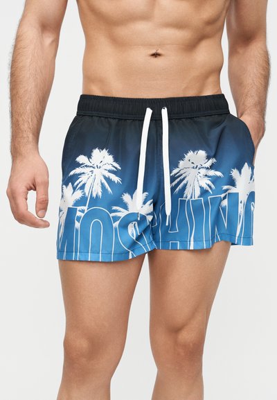 Torso masculin portant un short de bain bleu avec un imprimé de palmiers blancs et de grandes lettres partielles, mains détendues le long du corps.
