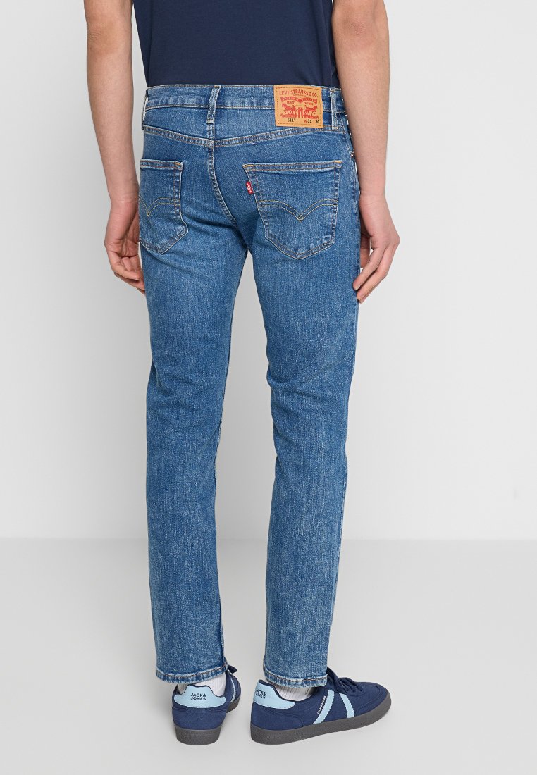 Vaqueros de mezclilla azul con un diseño de pierna recta, parche de cuero en la parte trasera y bolsillos traseros con un patrón de costura distintivo. Llevados con zapatillas náuticas.
