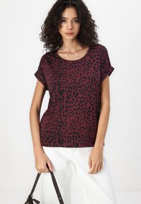 T-shirt imprimé léopard en bordeaux et noir, à manches courtes, encolure ronde, tissu doux, associé à un pantalon blanc et un sac à bandoulière noir.