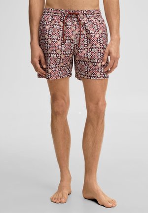 Man draagt een badshort met patroon en geometrische rode, roze en bruine vormen, staat blootsvoets tegen een lichtgrijze achtergrond.