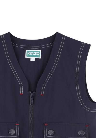 Marineblauwe mouwloze vest met een ritssluiting, contrasterende witte en rode stiksels, twee voorzakken met drukknoopsluitingen en een ronde halslijn.