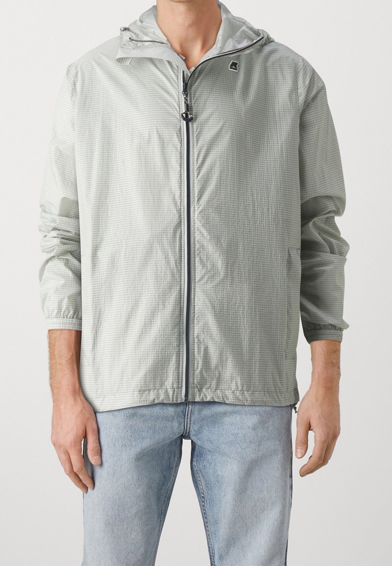Homme portant une veste à capuche gris clair avec un motif quadrillé subtil et un jean bleu clair, debout les bras détendus le long du corps.