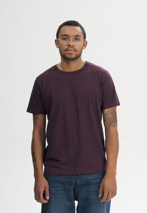 Man met kort haar en bril, draagt een bordeauxrood T-shirt en blauwe spijkerbroek, staand tegen een effen lichte achtergrond.