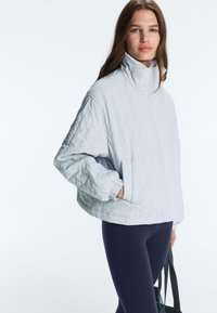 Nő hosszú barna hajjal, világosszürke, magas nyakú steppelt dzsekit visel és sötétkék leggingset, sötét színű táskát tart a kezében.