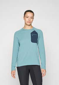 Langärmliges Funktionsshirt in Petrol mit einer blauen Tasche. Merkmale sind ein strukturiertes Gewebe und ein runder Ausschnitt. Logo-Detail auf der Tasche.